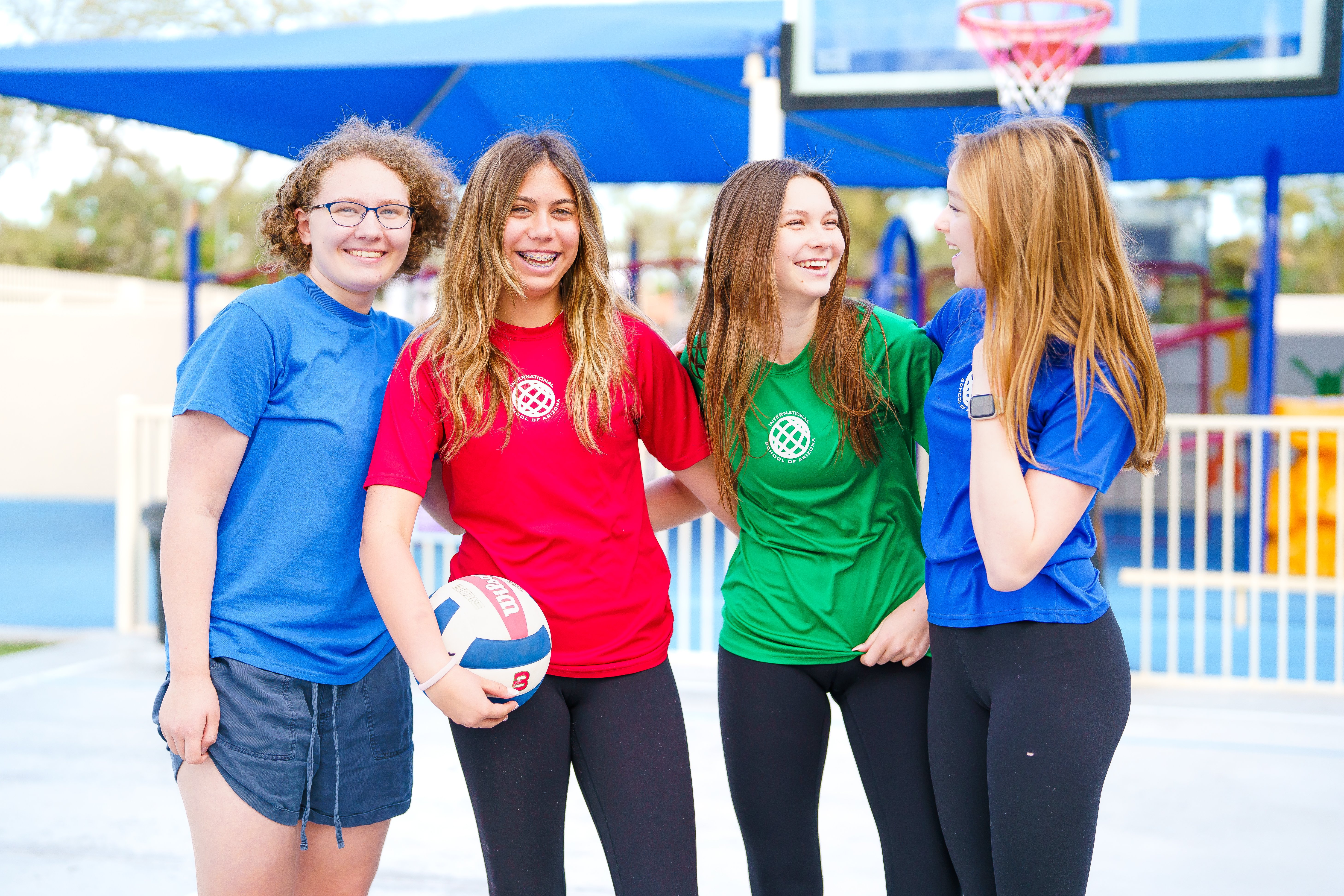  Group of middle school girls in sports uniforms smiling together with a volleyball at the International School of Arizona playground.